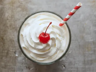 Concept imagery: a vanilla milkshake seen from above on a stainless counter, whipped cream with a cherry and a striped straw